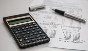 A close-up shows a black calculator with gold accents resting on papers filled with financial data and a pen.