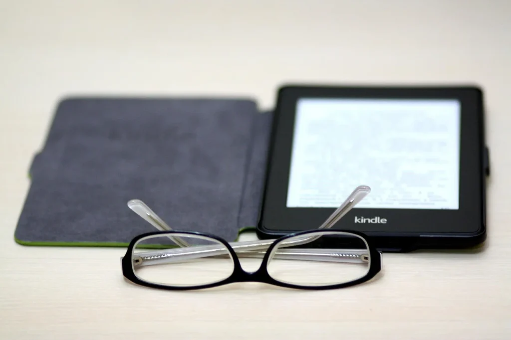 A Kindle e-reader in a black case with its screen lit up, resting on a light-colored surface next to a pair of black-framed reading glasses.