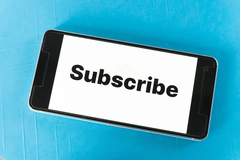 A close-up of a smartphone screen displaying the word "Subscribe" on a white background, on a blue surface.